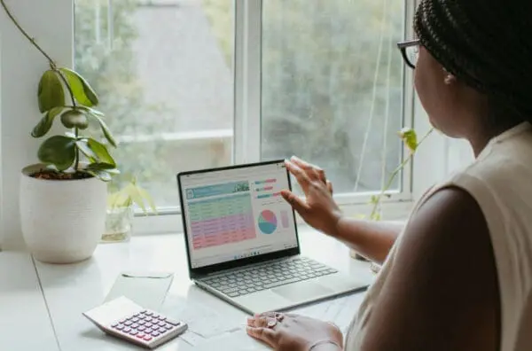 A person wearing glasses and braided hair is working on a laptop at a white desk near a window. The laptop screen displays colorful charts and graphs, possibly for an SEO blog. A plant, a calculator, and some papers are on the desk. The background shows blurred greenery outside the window.